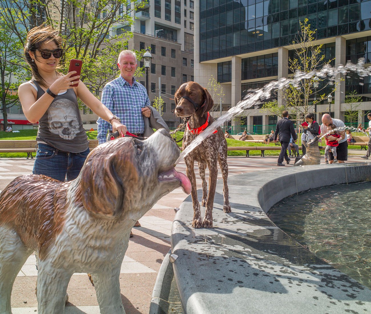 A young woman taking a picture of the fountain at Berczy Park in Toronto while her dog is looking at the statue